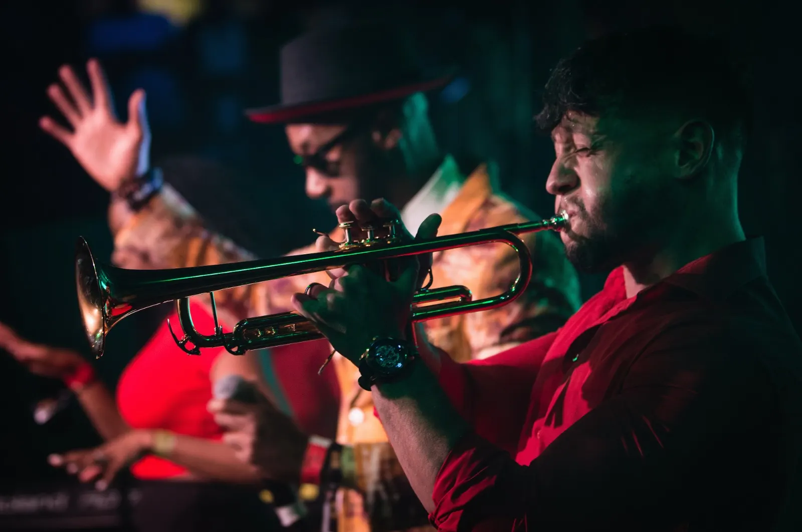 Vox Sambou with his hand raised behind the trumpet player, the band lit in red and green